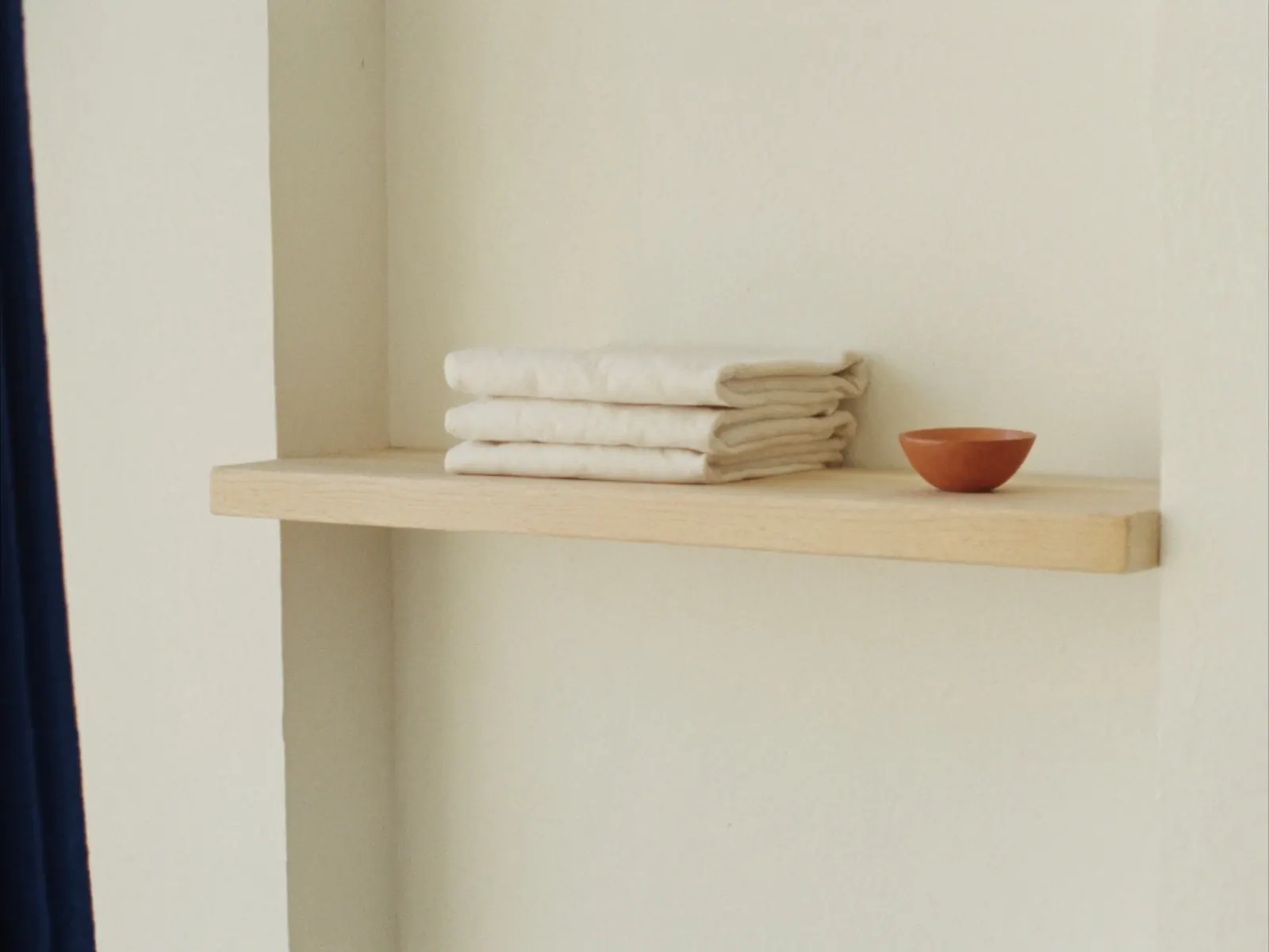 A pale oak wall shelf holding three folded linen towels and a small terracotta bowl, in soft daylight
