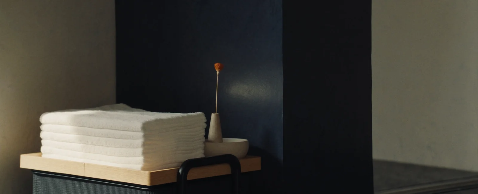 Folded white towels on a pale oak trolley top beside a stone bud vase and a ceramic bowl, against a deep navy wall