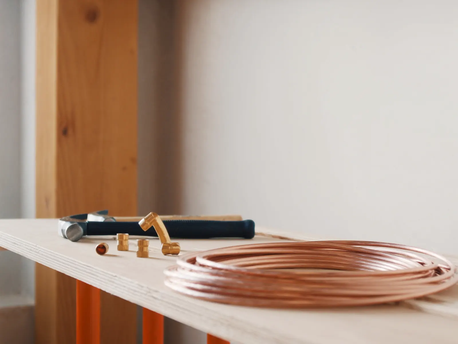 A coil of copper pipe, brass fittings and a hammer on a pale wooden bench in soft daylight